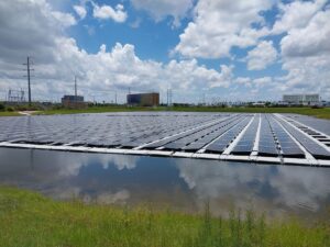 A large array of floating Epic Universe solar panels on a calm pond under a bright blue sky with white clouds, with power lines and distant buildings in view.