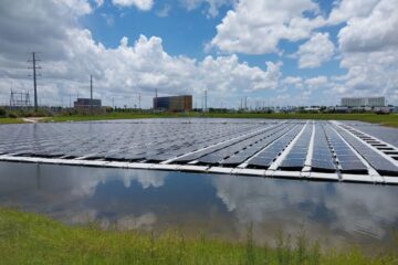 A large array of floating Epic Universe solar panels on a calm pond under a bright blue sky with white clouds, with power lines and distant buildings in view.