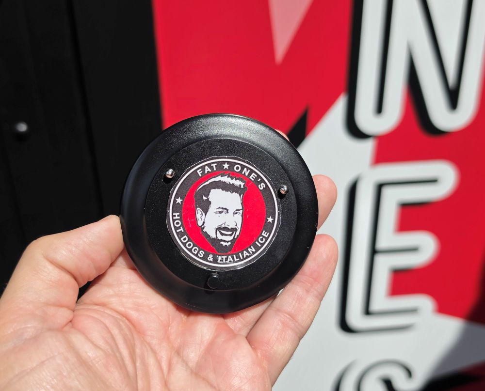 A hand holds a black circular restaurant pager featuring the Fat One's Hot Dogs & Italian Ice logo with a man's face against a red background.