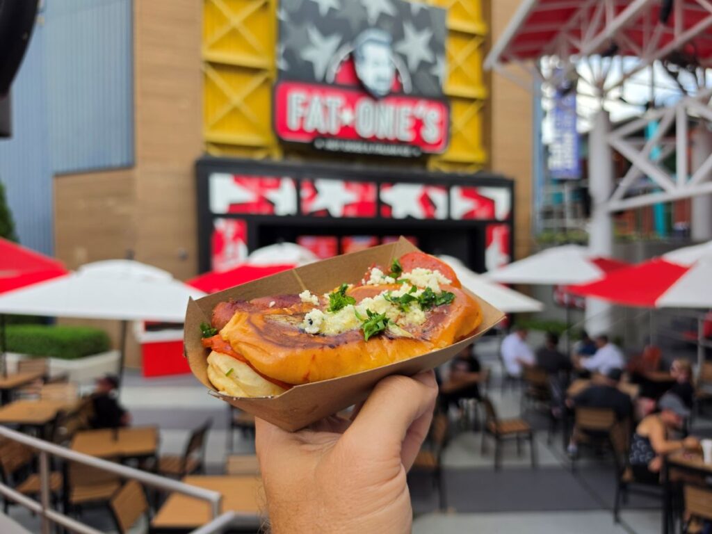 A hand holds a hot dog in a cardboard tray, topped with cheese and herbs, with a blurred Fat One's restaurant and outdoor seating in the background.
