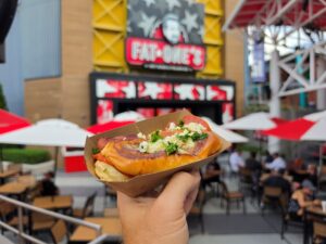 A hand holds a hot dog in a cardboard tray, topped with cheese and herbs, with a blurred Fat One's restaurant and outdoor seating in the background.