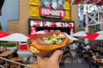 A hand holds a hot dog in a cardboard tray, topped with cheese and herbs, with a blurred Fat One's restaurant and outdoor seating in the background.