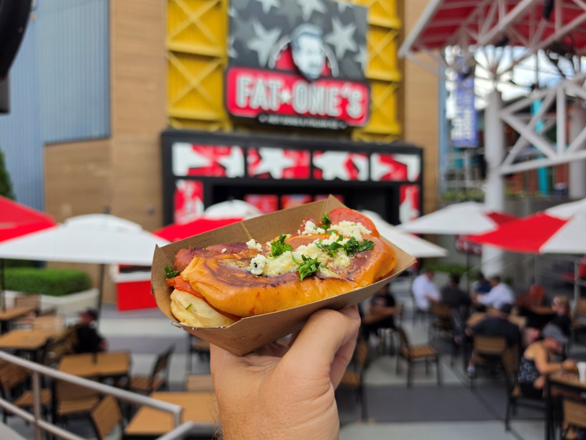 A hand holds a hot dog in a cardboard tray, topped with cheese and herbs, with a blurred Fat One's restaurant and outdoor seating in the background.