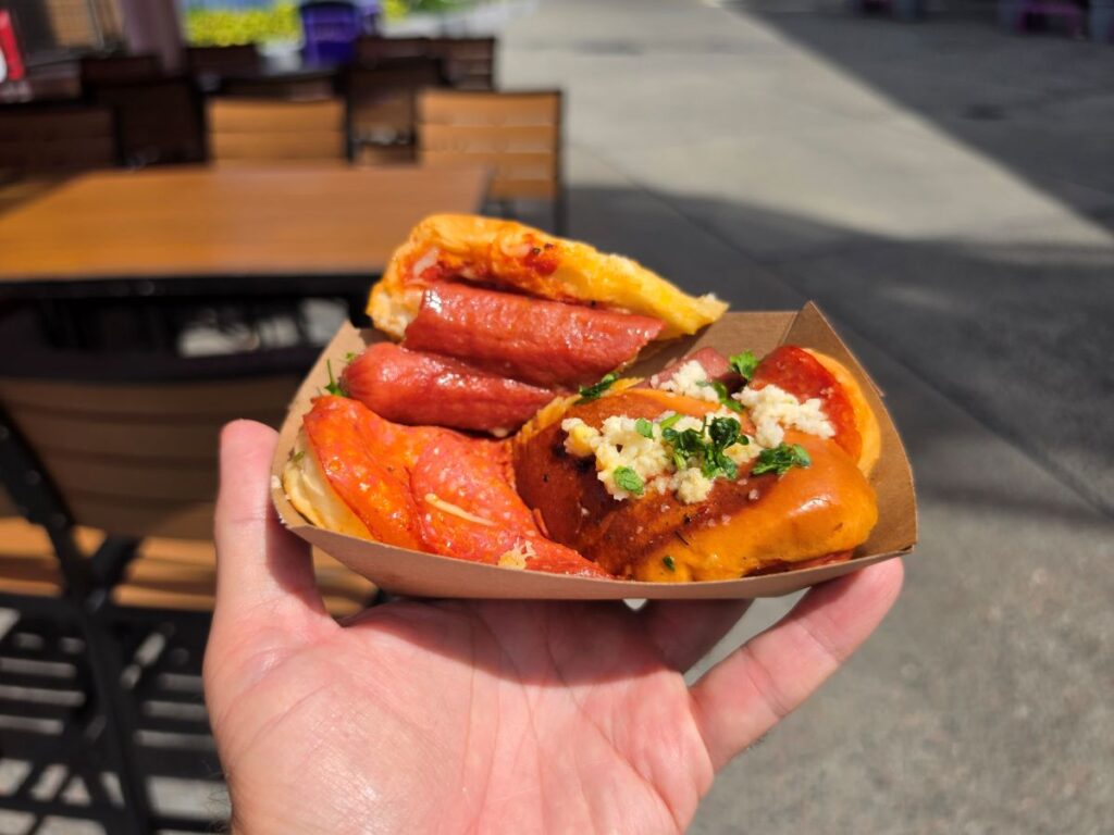 A hand holds a cardboard tray containing two small, savory pastries topped with sausage, pepperoni, melted cheese, and fresh green herbs outdoors.