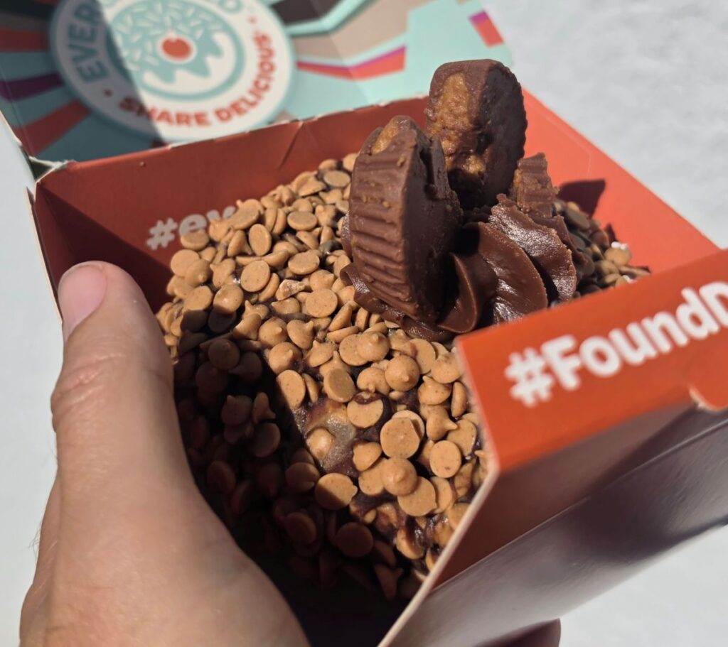 A glazed donut covered in small peanut butter chips, topped with chocolate frosting and a broken peanut butter cup, held in an orange cardboard box.