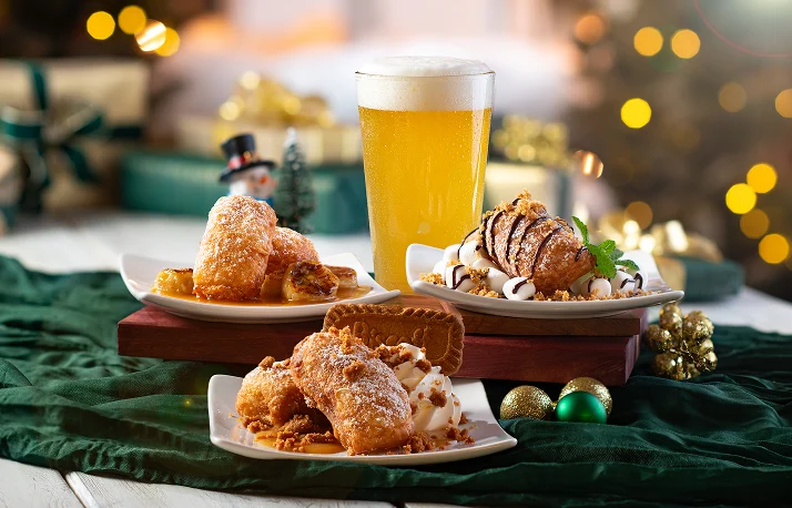 Three plates of fried pastries with toppings, a tall glass of golden beer, and holiday decorations on a green cloth against a blurred bokeh background.