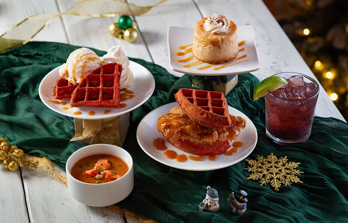 A festive holiday spread featuring red waffles with ice cream, a small cake, fried chicken on a waffle, lobster bisque, and a dark cocktail drink.
