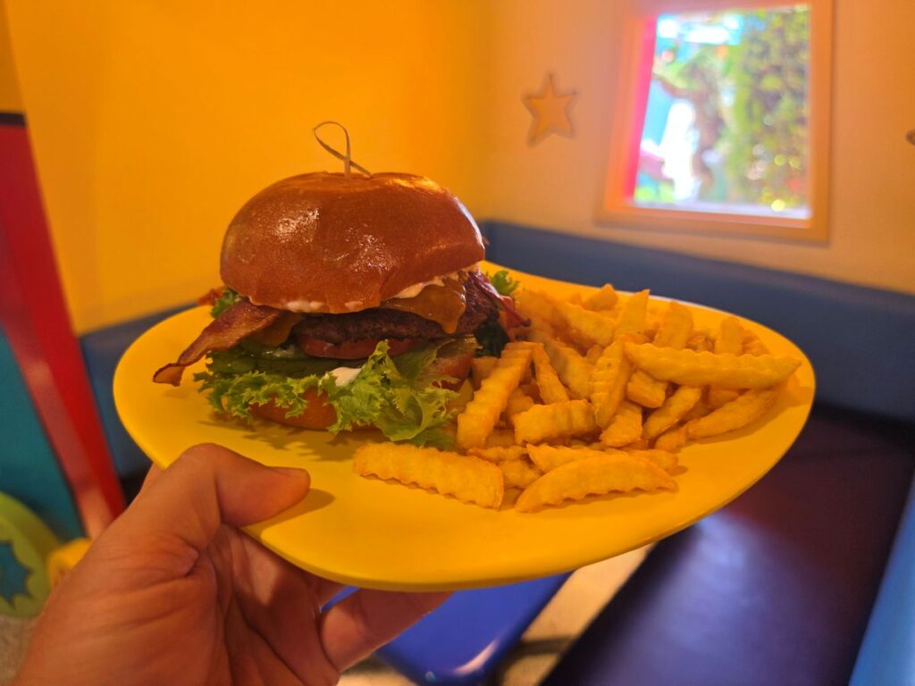 A hand holds a yellow plate with a Roast Beast bacon cheeseburger and crinkle-cut fries in a brightly colored, toy-themed room with a star on the yellow wall.