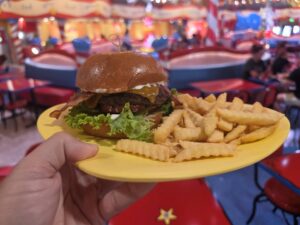 A hand holds a yellow plate with a Roast Beast bacon cheeseburger and crinkle-cut fries inside a brightly colored, whimsical restaurant with red and blue decor.