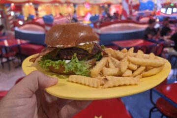 A hand holds a yellow plate with a Roast Beast bacon cheeseburger and crinkle-cut fries inside a brightly colored, whimsical restaurant with red and blue decor.