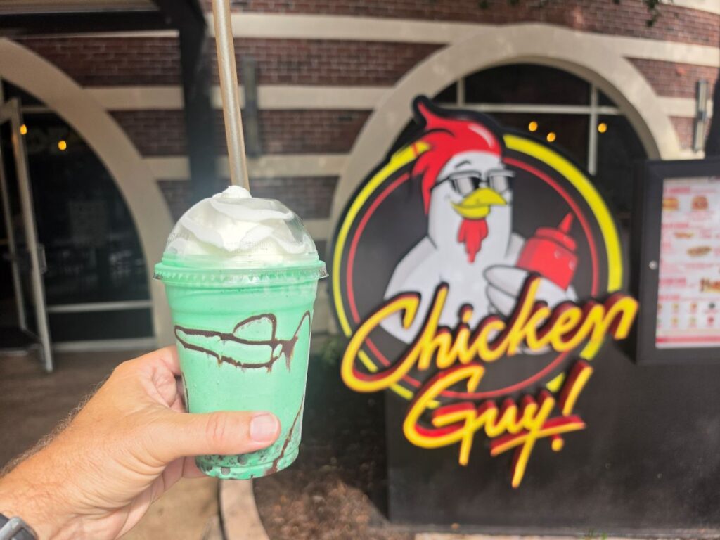 A hand holds a mint green milkshake with whipped cream and chocolate drizzle in front of a Chicken Guy! restaurant sign featuring a cartoon chicken.