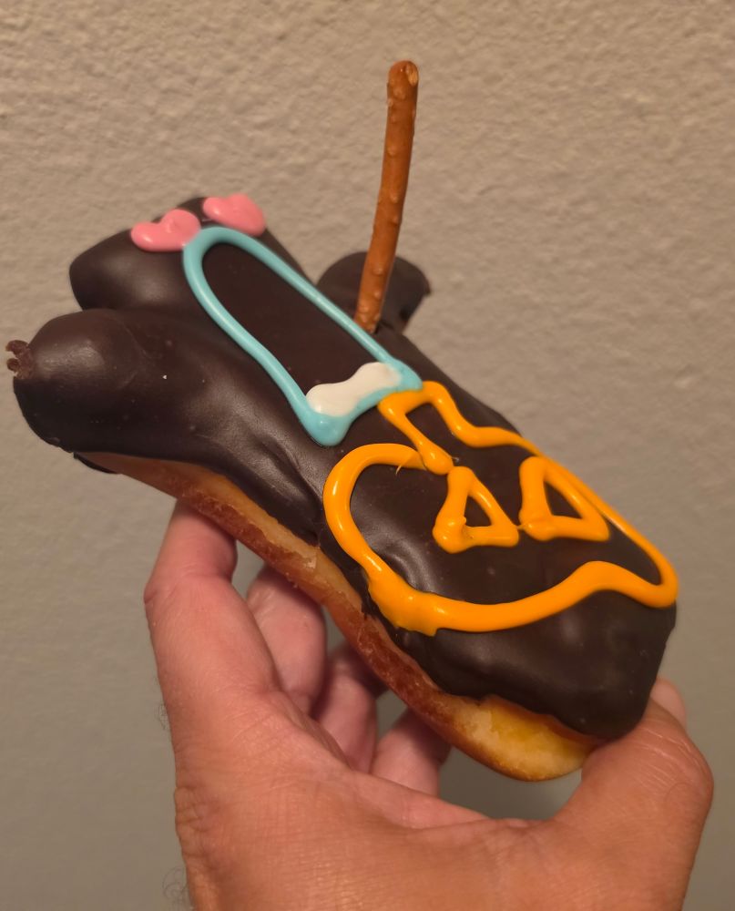 A hand holds a chocolate-glazed eclair decorated with orange icing, a blue outline, pink heart sprinkles, and a single vertical pretzel stick.