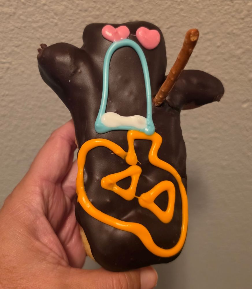 A hand holds a chocolate-covered donut shaped like a voodoo doll, decorated with pink heart eyes, a blue bow tie, an orange pumpkin, and a pretzel stick.