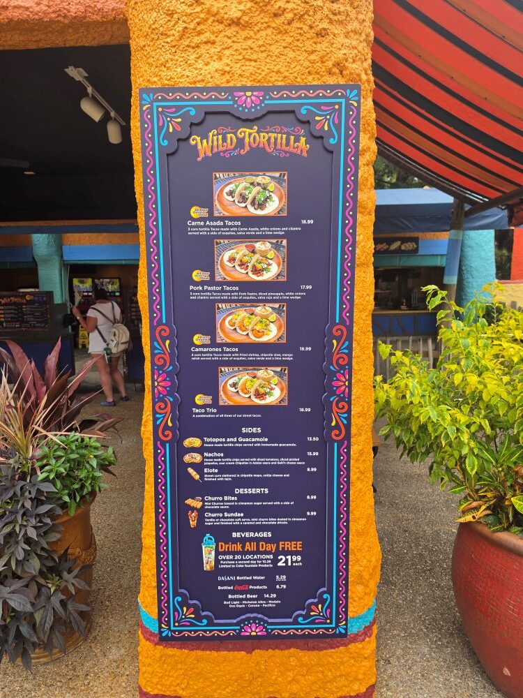 A vertical menu board for Wild Tortilla mounted on an orange textured pillar, listing various taco options, sides, desserts, and beverage prices.